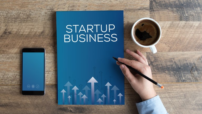 a mobile phone on a table next to a book titled business startup with a hand holding a pen and a cup of coffee