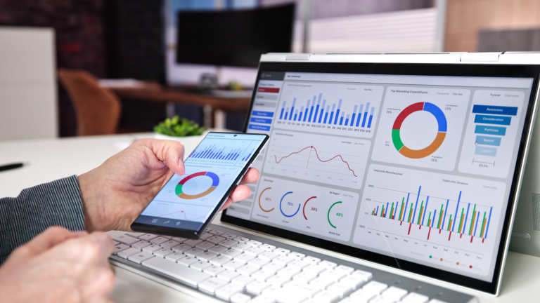 person holding a smartphone with laptop in the background showing website analytics